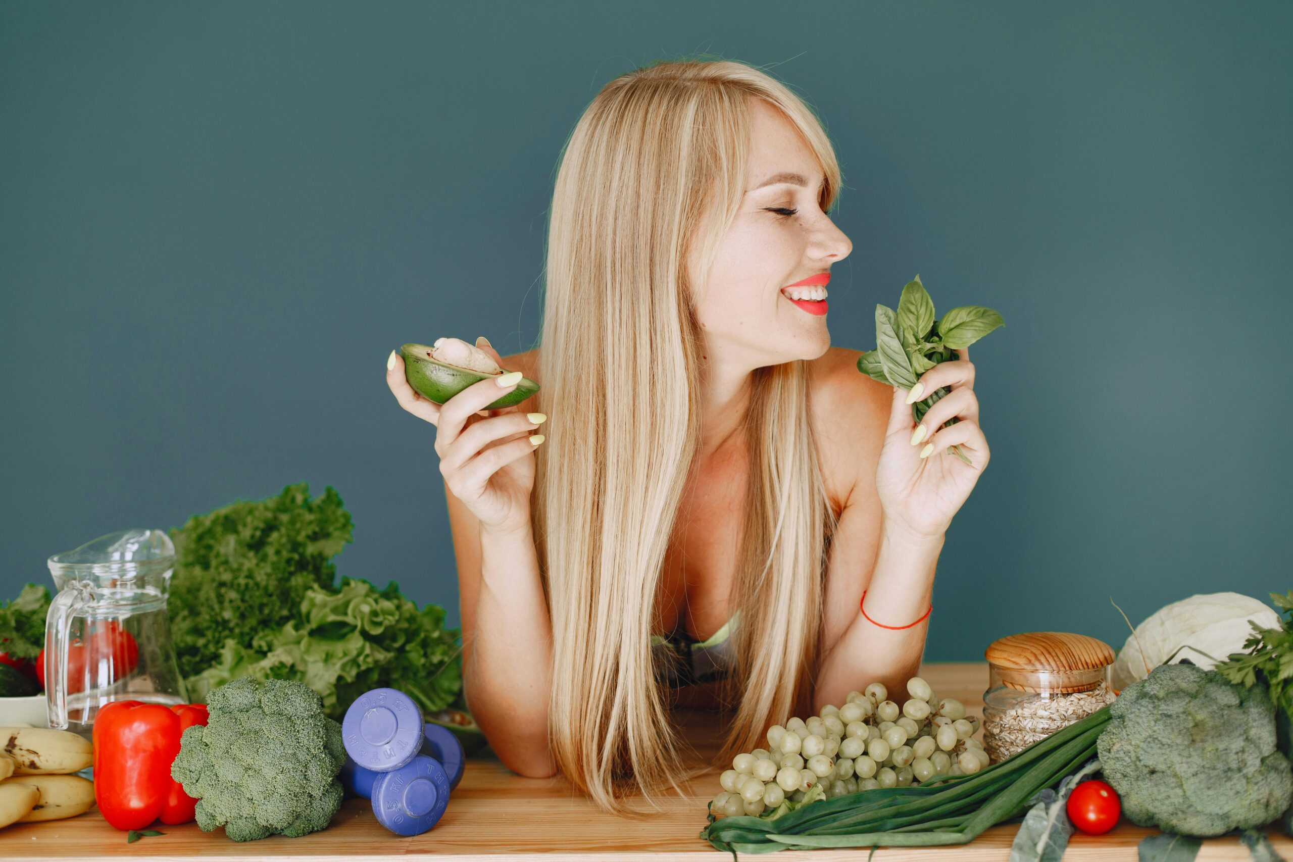 A smiling woman holding avocado and basil, surrounded by fresh vegetables and fruits for healthy eating.